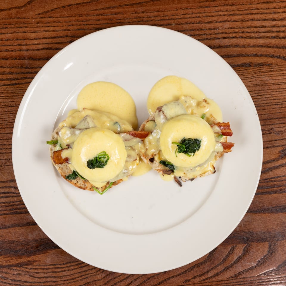 Two servings of Eggs Benedict with poached eggs, hollandaise, spinach, and bacon on a white plate.