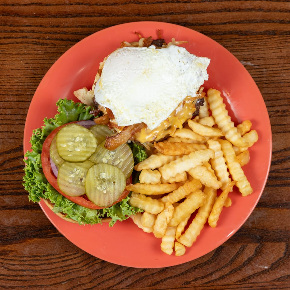 Burger with fried egg, cheese, bacon, lettuce, tomato, pickles, and crinkle-cut fries on an orange plate.