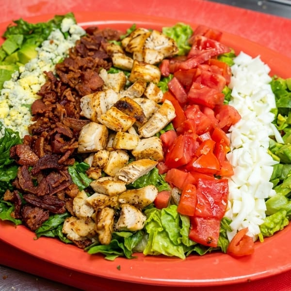 Vibrant Cobb salad on a red plate, featuring rows of lettuce, avocado, blue cheese, bacon, chicken, tomatoes, and egg.