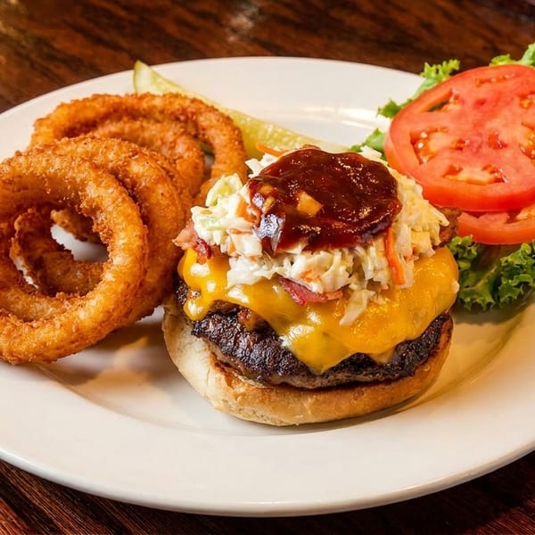 A cheeseburger with coleslaw and BBQ sauce, served with crispy onion rings, a pickle, lettuce, and tomato.